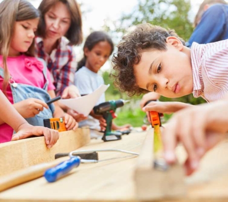 Kinder handwerken auf einem Tisch, Bub misst etwas und zwei Mädchen und eine Frau schauen auf die Arbeitsplatte