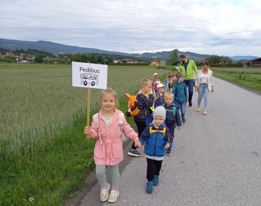 Kinder gehen Hand in Hand auf einer Straße, die von Feldern begrenzt ist, das erste Kind hält ein Pedibus-Schild in der Hand