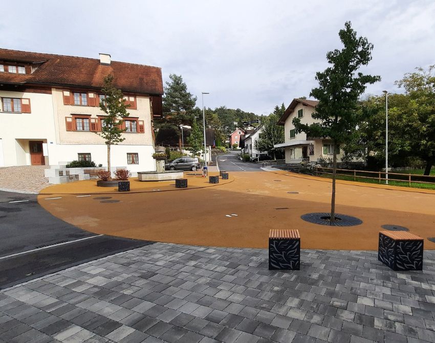 Dorfplatz mit einem jungen Baum in der Mitte und ockerfarbenem Asphalt, rundherum eine Pflasterung