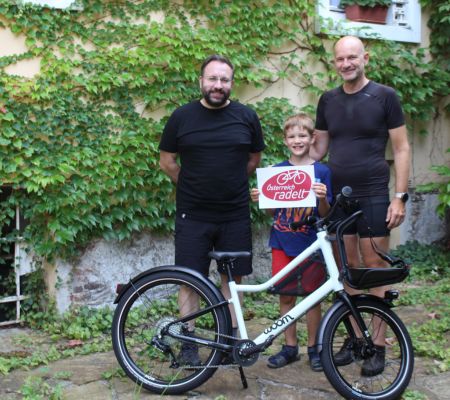 2 Männer, 1 Kind stehen hinter einem Kinderfahrrad, das Kind hat das Österreich radelt Logo in A4-Größe in der Hand