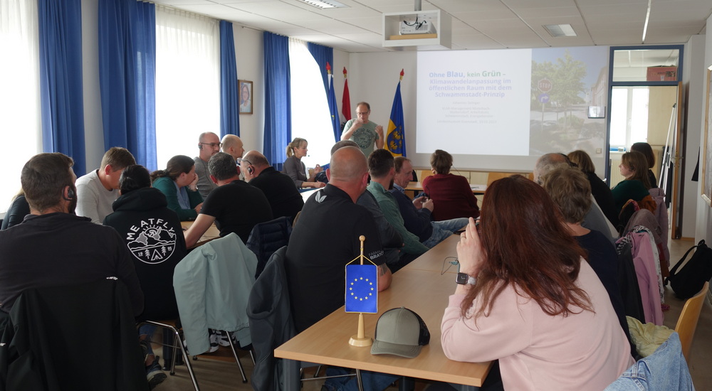 Die Gruppe sitzt in einem Saal und härt einem Vortrag zu. Im Vordergrund steht eine EU Flagge.