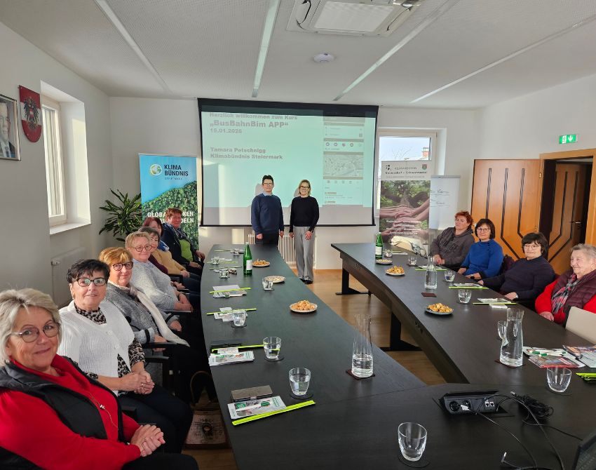 Senioren sitzen an langer Tafel und lächeln, im Hintergrund Beamer und Vortragende