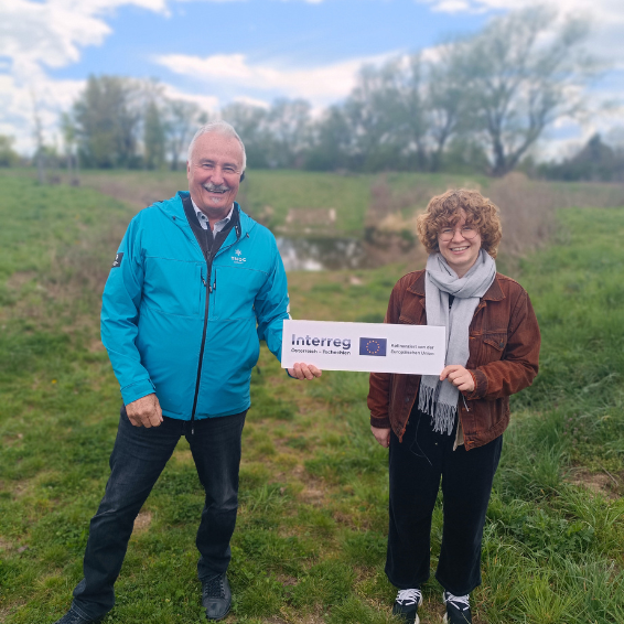 Der Umweltstadtrat und eine Mitarbeiterin des Klimabündnis lachen in die Kamera und halten ein Interreg Schild in die Höhe