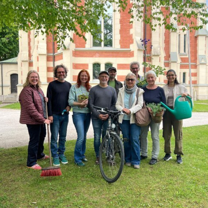 Gruppenfoto von ungefähr 10 Personen. Sie halten Pflanzen, Besen, eine Gießkanne und ein Fahrrad