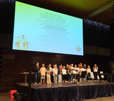 Schüer:innen der Volksschule St. Anton am Arlberg singen den Klimasong auf einer Bühne, im Hintergrund der Text des Liedes